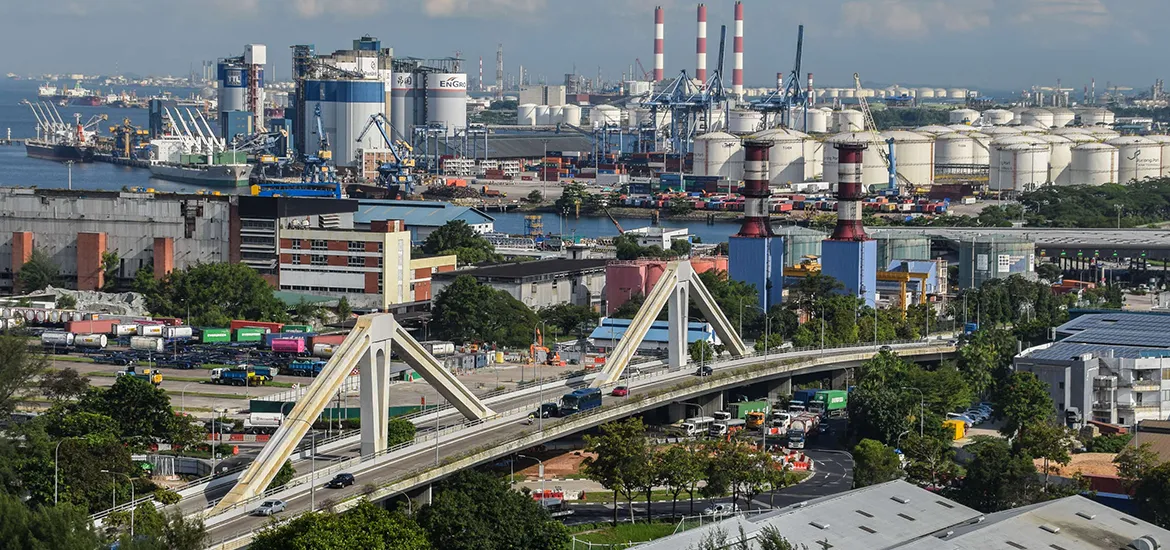 View of Singapore’s industrial port area with factories, storage tanks, cranes, and a highway bridge in the foreground.