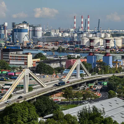 View of Singapore’s industrial port area with factories, storage tanks, cranes, and a highway bridge in the foreground.