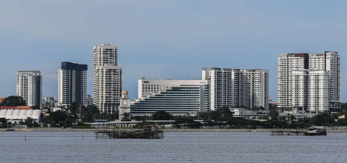 Cityscape of Johor Bahru with modern high-rise buildings and waterfront view under a clear blue sky.