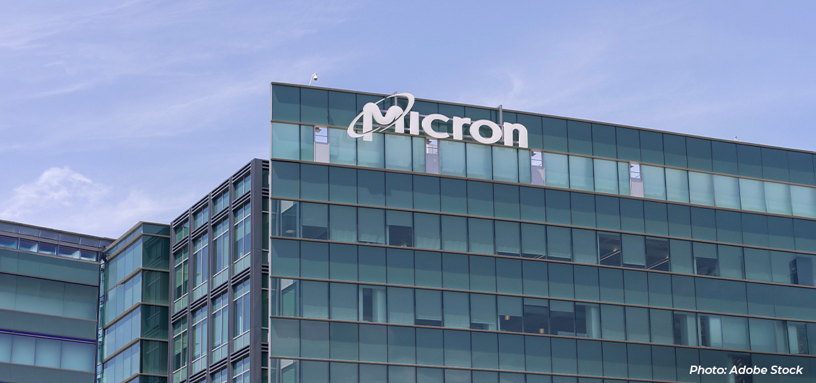 Exterior view of a modern Micron office and manufacturing building with a glass façade and the Micron logo prominently displayed on the upper section of the structure against a cloudy blue sky. Photo credit: Adobe Stock.