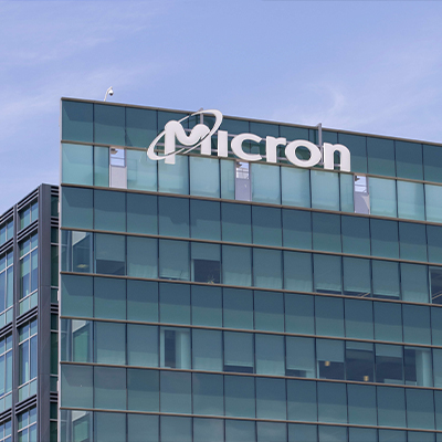 Exterior view of a modern Micron office and manufacturing building with a glass façade and the Micron logo prominently displayed on the upper section of the structure against a cloudy blue sky.