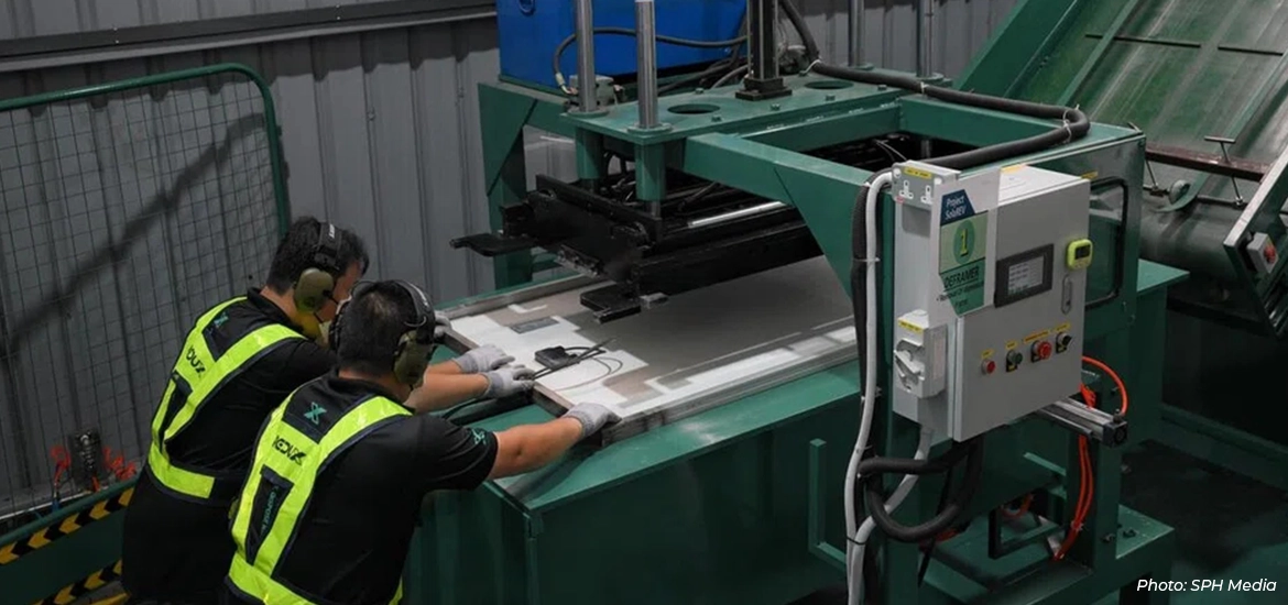 Two factory workers operate industrial machinery on a manufacturing line, handling flat metal sheets in a production facility.