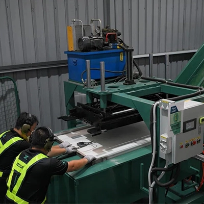 Two factory workers operate industrial machinery on a manufacturing line, handling flat metal sheets in a production facility.