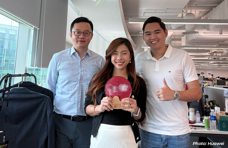 Three Huawei employees posing together in a modern office workspace, with one employee holding a commemorative gift while colleagues smile and gesture towards the camera, highlighting workplace recognition and team spirit.