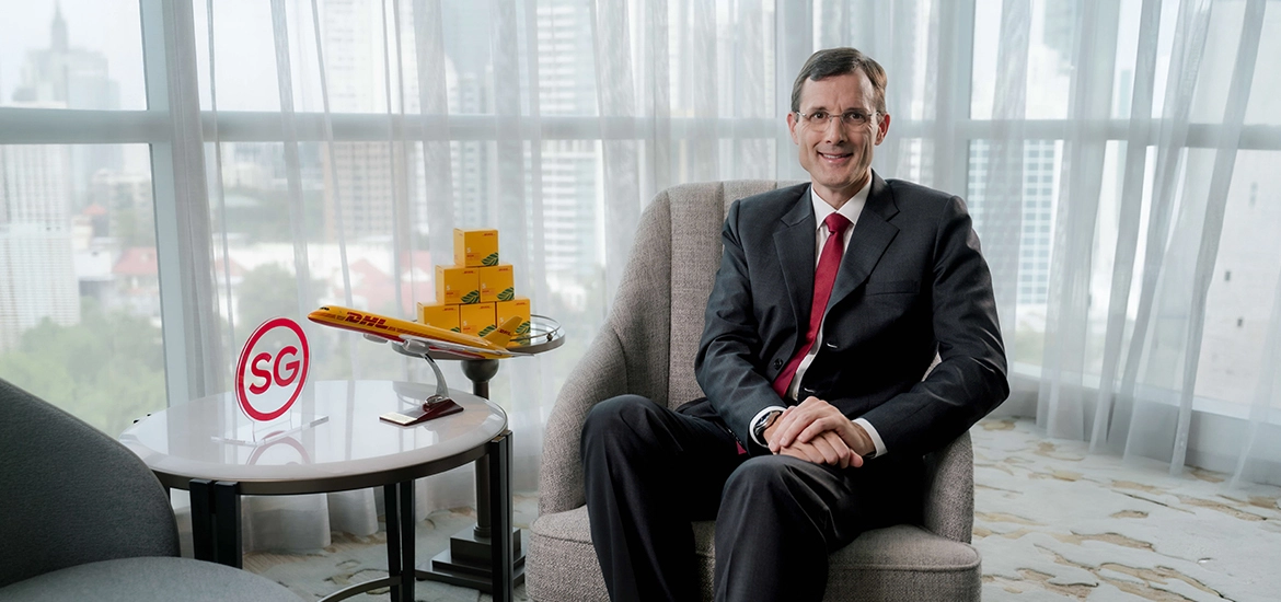 Tobias Meyer seated in a corporate setting, with a DHL model airplane and boxes on a side table alongside an “SG” sign, representing DHL’s Asia Pacific presence.