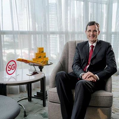 Tobias Meyer seated in a corporate setting, with a DHL model airplane and boxes on a side table alongside an “SG” sign, representing DHL’s Asia Pacific presence.