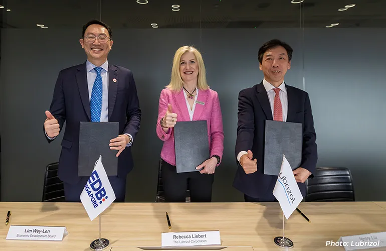 Three executives holding signed documents and giving thumbs up at a partnership signing, with EDB Singapore and Lubrizol flags on the table.