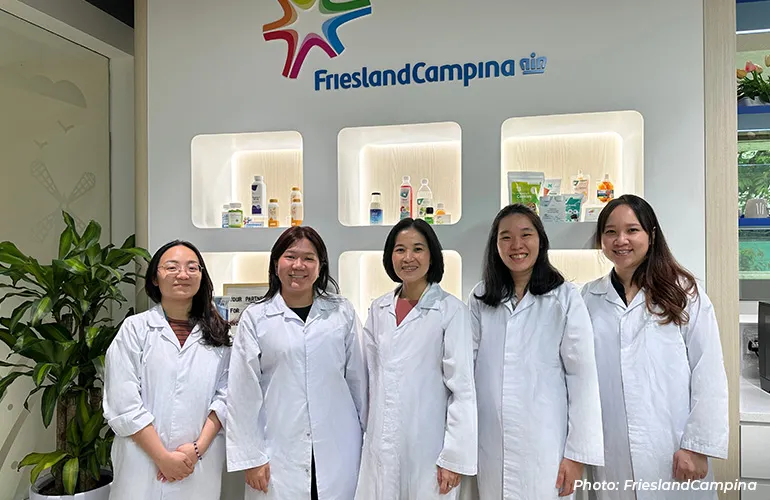 Five researchers in white lab coats standing in front of a FrieslandCampina display with product samples on shelves.
