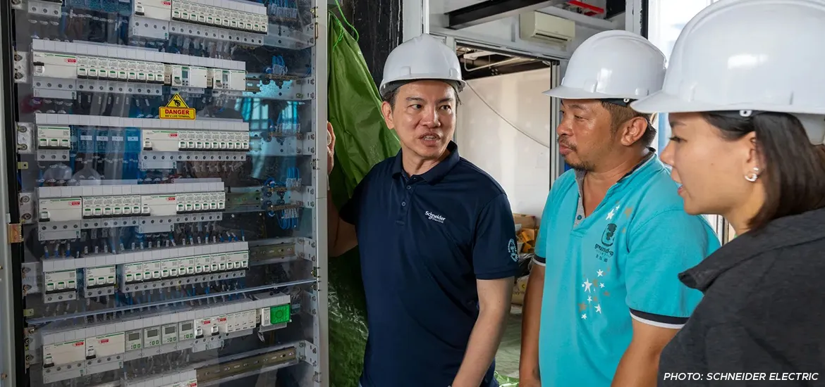 Three engineers wearing safety helmets inspecting an electrical control panel during a discussion at a worksite.