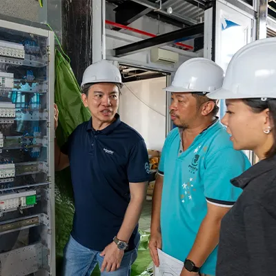 Three engineers wearing safety helmets inspecting an electrical control panel during a discussion at a worksite.