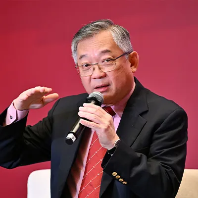 Man in a suit speaking into a microphone during a discussion, gesturing with his hand against a red background.