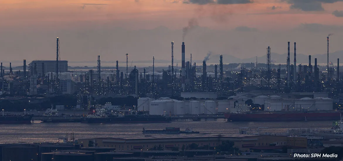 Industrial refinery and petrochemical complex at dusk, with storage tanks, ships, and smokestacks along the waterfront.