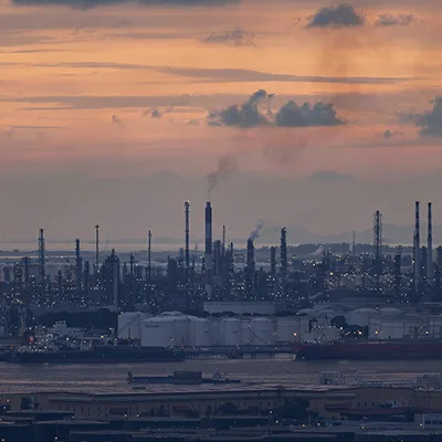 Industrial refinery and petrochemical complex at dusk, with storage tanks, ships, and smokestacks along the waterfront.