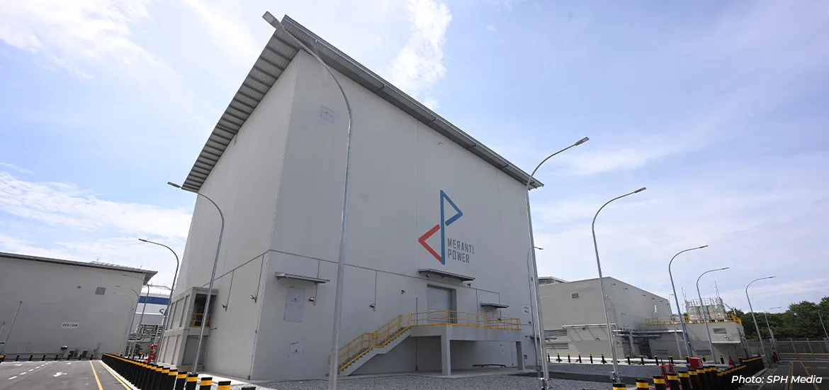 Exterior view of a modern Meranti Power industrial facility in Singapore, featuring a large grey energy building with company logo, safety railings, street lighting, and surrounding utility infrastructure under a clear sky.