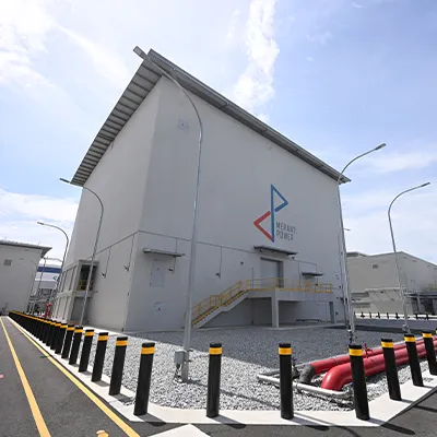 Exterior view of a modern Meranti Power industrial facility in Singapore, featuring a large grey energy building with company logo, safety railings, street lighting, and surrounding utility infrastructure under a clear sky.