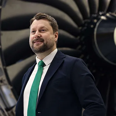 Man in a suit with a green tie standing in front of a large aircraft jet engine.