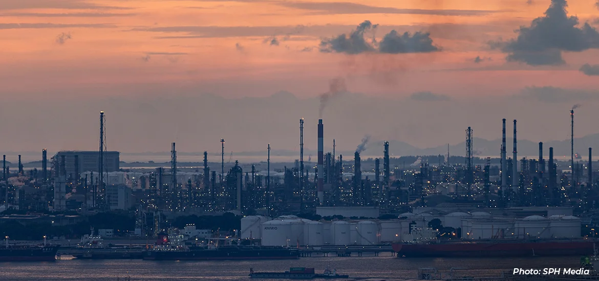 Petrochemical and refinery facilities on Jurong Island at sunset, with storage tanks, industrial towers, smokestacks, and ships along the waterfront under a pink-orange sky.