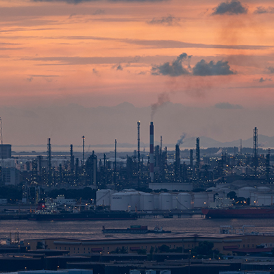 Petrochemical and refinery facilities on Jurong Island at sunset, with storage tanks, industrial towers, smokestacks, and ships along the waterfront under a pink-orange sky.