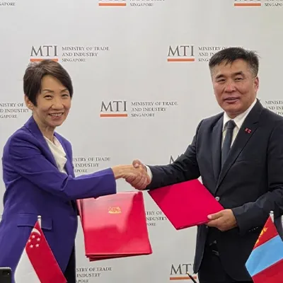 Representatives from Singapore’s Ministry of Trade and Industry and another country shaking hands after signing an agreement, holding red folders in front of an MTI backdrop.