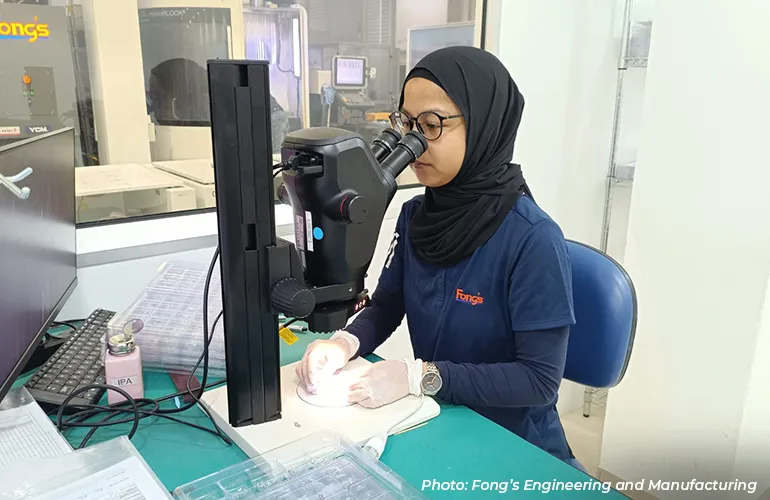 Technician wearing gloves and a headscarf examining a component through a microscope in a laboratory workspace.