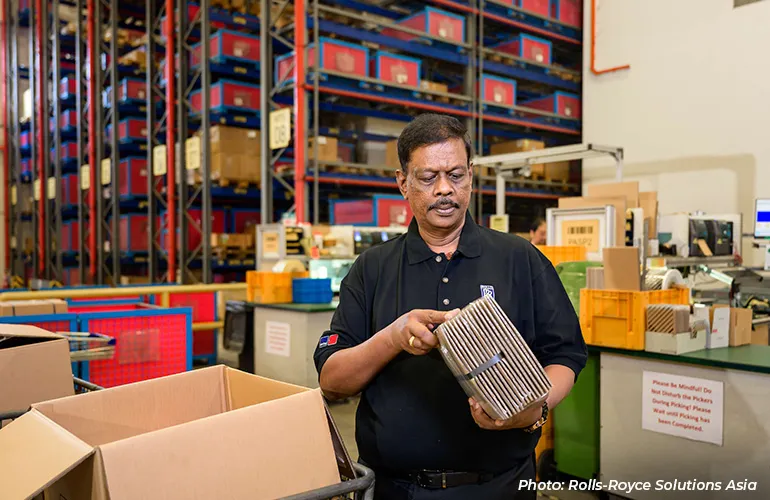 Worker inspecting a metal component inside a warehouse with shelves of parts and boxes in the background.