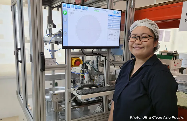 Technician wearing a hairnet and glasses standing beside automated manufacturing equipment and a monitor display.