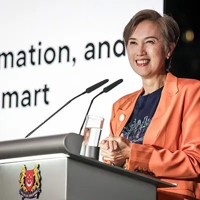 Smiling woman speaking at a podium during a conference, with presentation text in the background.