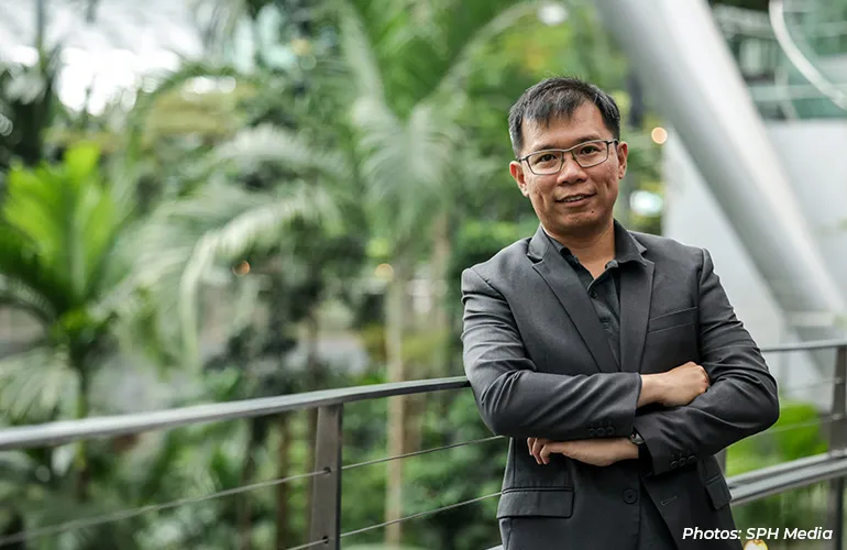A man in a dark blazer stands with arms crossed in a lush indoor garden setting.