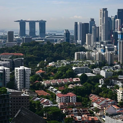 Aerial view of Singapore's urban landscape featuring residential neighborhoods in the foreground and the iconic Marina Bay Sands hotel and downtown skyscrapers in the background, surrounded by lush greenery under a clear blue sky.
