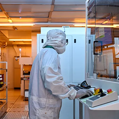Technician in a cleanroom suit operating semiconductor manufacturing equipment inside a fabrication facility.