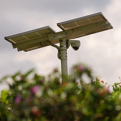 Solar-powered CCTV camera mounted on a pole above greenery, with cloudy sky in the background.