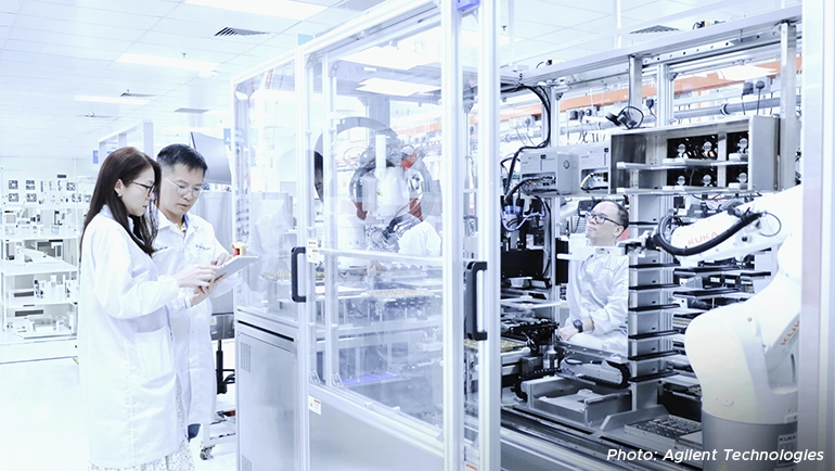 Researchers in lab coats working with advanced automated laboratory equipment in a cleanroom setting at Agilent Technologies.