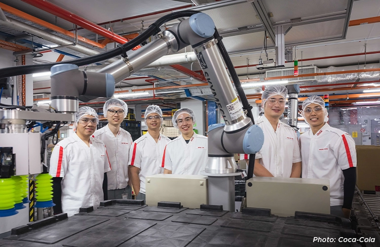 A group of Coca-Cola employees in lab coats, hairnets, and safety glasses standing beside an industrial robotic arm inside a modern production facility.