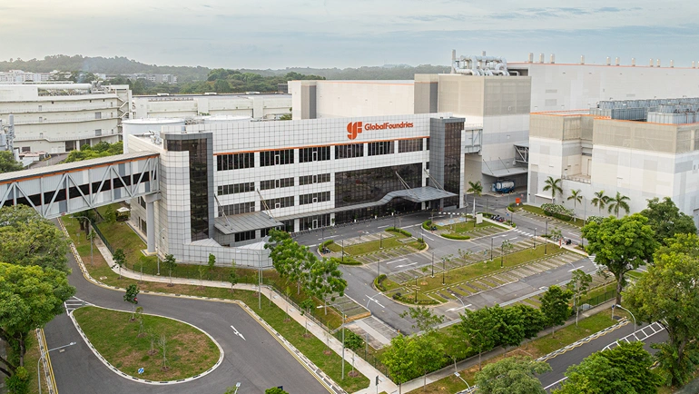 Aerial view of the GlobalFoundries semiconductor manufacturing facility in Singapore, featuring large modern factory buildings surrounded by greenery and roads.