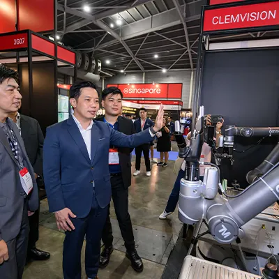 Group of professionals observing a demonstration of robotic arms at a technology exhibition, with "Singapore" signage visible in the background.