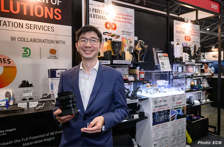 Man in a suit smiling and holding a 3D-printed component at a booth showcasing 3D printing solutions during a trade exhibition.