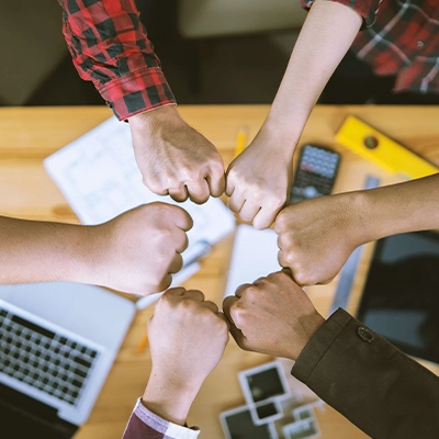 A top-down view of six diverse hands coming together in a fist bump over a wooden table, symbolizing teamwork and unity. Office items such as a laptop, tablet, calculator, ruler, and papers are scattered around, indicating a collaborative work environment.