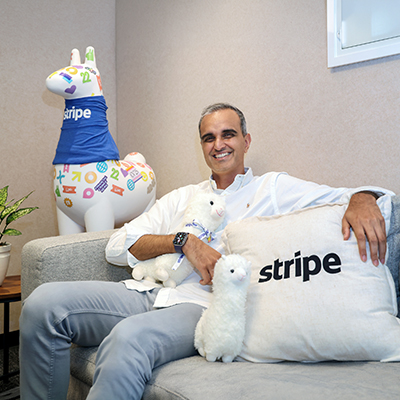 Portrait of a Stripe executive seated in a lounge area with branded décor and plush mascots, smiling in a relaxed office environment that reflects the company's workplace culture.