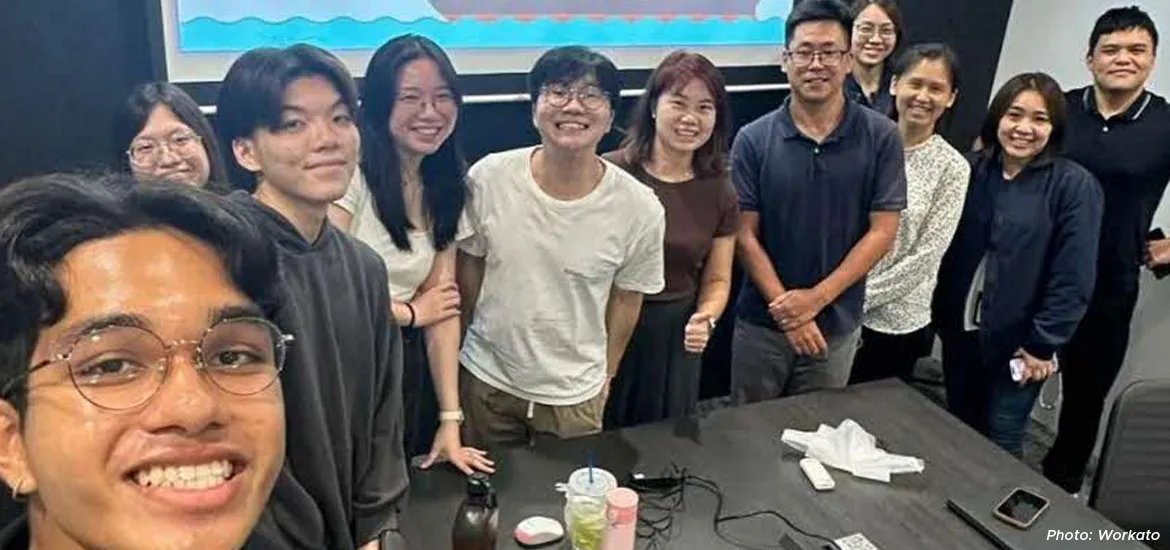 Group of young professionals smiling and posing together in a meeting room with a presentation screen behind them.