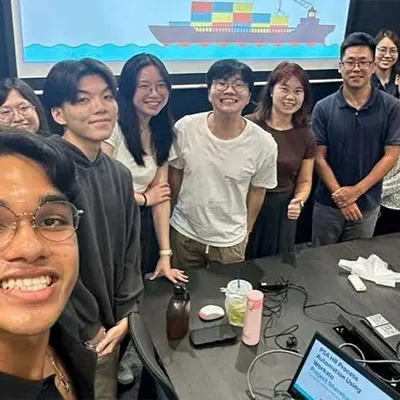Group of young professionals smiling and posing together in a meeting room with a presentation screen behind them.