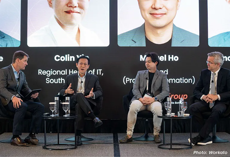 Panel discussion at a conference featuring four speakers seated on stage, engaged in conversation in front of a large screen displaying their names and titles.