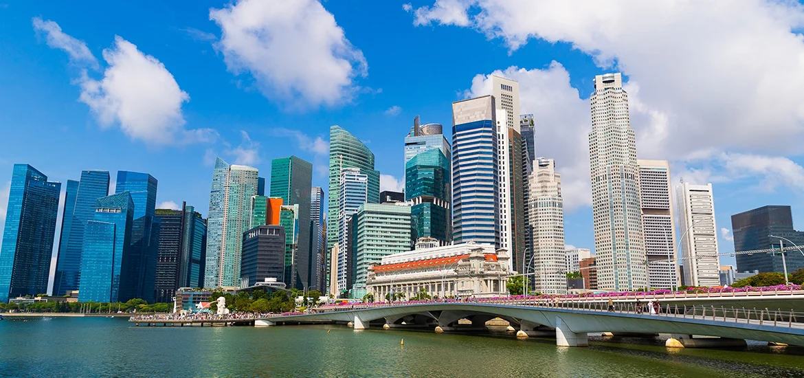 Singapore city skyline with high-rise buildings and a bridge over Marina Bay under a blue sky.