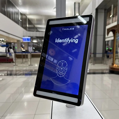 Tablet displaying a facial recognition identification screen in an airport terminal, with security screening area in the background.