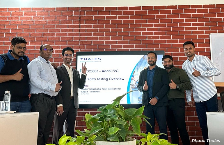 Group of professionals standing in an office, posing in front of a screen displaying a Thales project presentation, with several giving thumbs-up gestures.