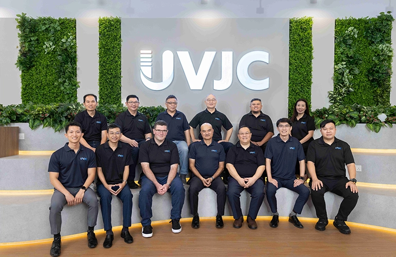 Group photo of the UVJC team seated and standing in an office space, with a large UVJC logo and greenery wall in the background.
