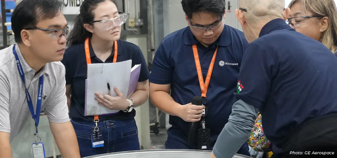 GE Aerospace engineers and technicians wearing safety glasses collaborate on the factory floor, reviewing a large aircraft component during hands-on training and advanced aerospace manufacturing operations.