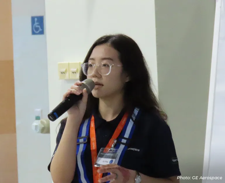 GE Aerospace employee wearing safety glasses and a company uniform speaks into a microphone during a workplace briefing or technical presentation, highlighting employee engagement and advanced aerospace operations.