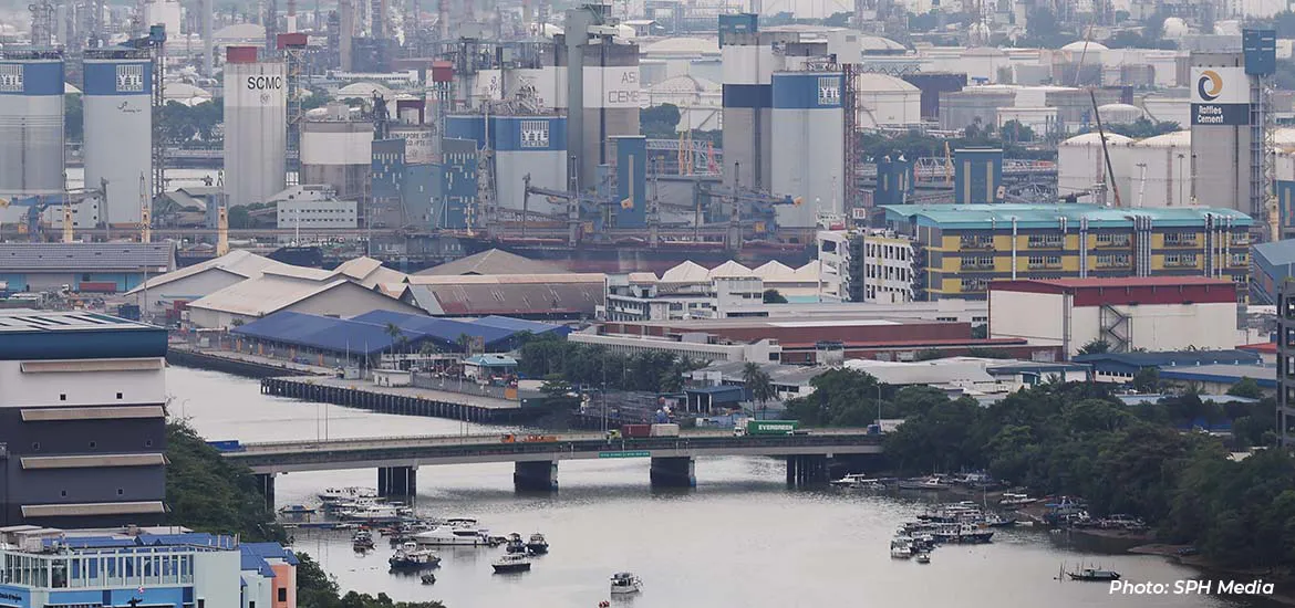 An industrial waterfront area with factories, storage tanks, and warehouses, with a bridge crossing a river where small boats are docked.