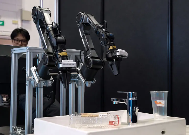 Two robotic arms mounted on a frame operating above a sink, with a faucet, plastic bottle, and cup on the counter while a person observes in the background.