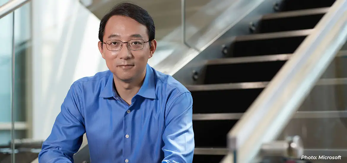 Man in a blue shirt and glasses seated in a modern office setting, with a staircase in the background.